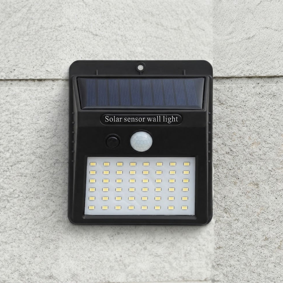 A black solar sensor wall light mounted on a textured light gray stone wall. The light features a dark blue solar panel at the top, a white motion sensor, and a grid of LED lights at the bottom, with the text 'Solar sensor wall light' printed on the front.
