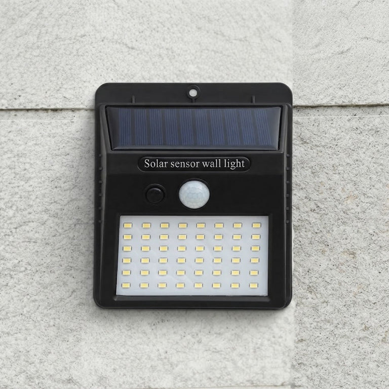 A black solar sensor wall light mounted on a textured light gray stone wall. The light features a dark blue solar panel at the top, a white motion sensor, and a grid of LED lights at the bottom, with the text 'Solar sensor wall light' printed on the front.